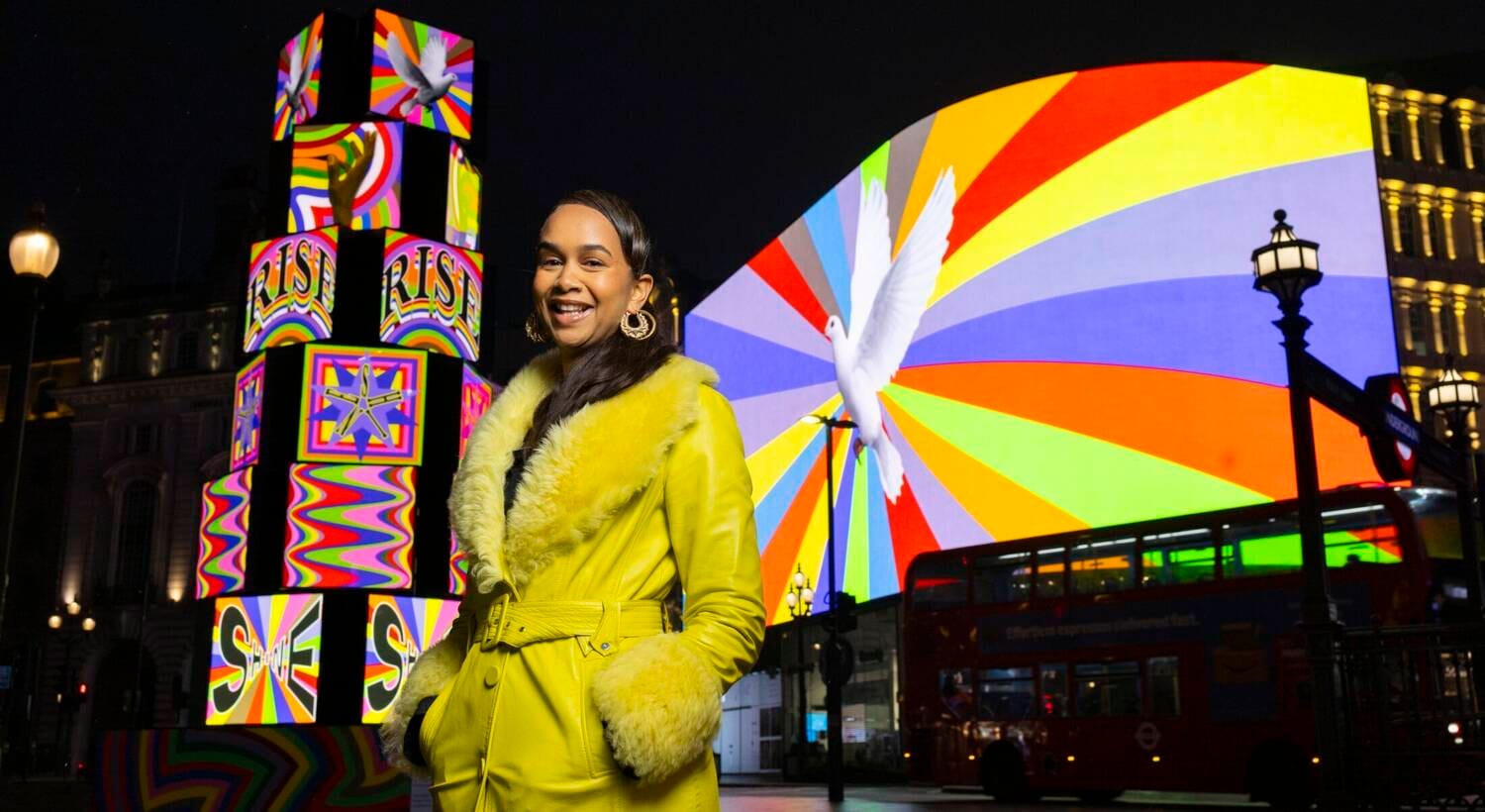 Lakwena Maciver in front of Rise and Shine, a 7-metre tower of stacked disco lightboxes at Piccadilly Circus, 2026